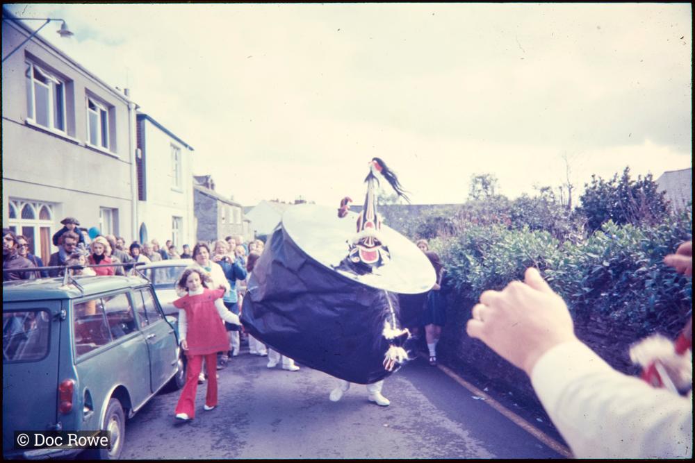 Old Oss walking along road, holding hands with girl, crowd behind
