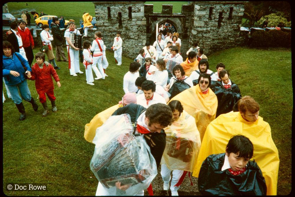 Old Oss procession accordion players wearing rain ponchos, walking through Prideaux Place gates